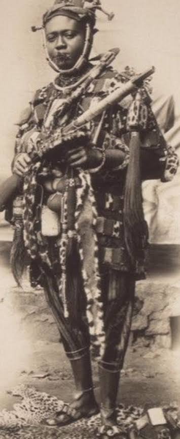The Women Who Built the Revolution 2 A historical black and white full-length portrait of Nana Yaa Asantewaa in traditional Ashanti warrior dress, standing with a rifle to represent her leadership in the War of the Golden Stool.