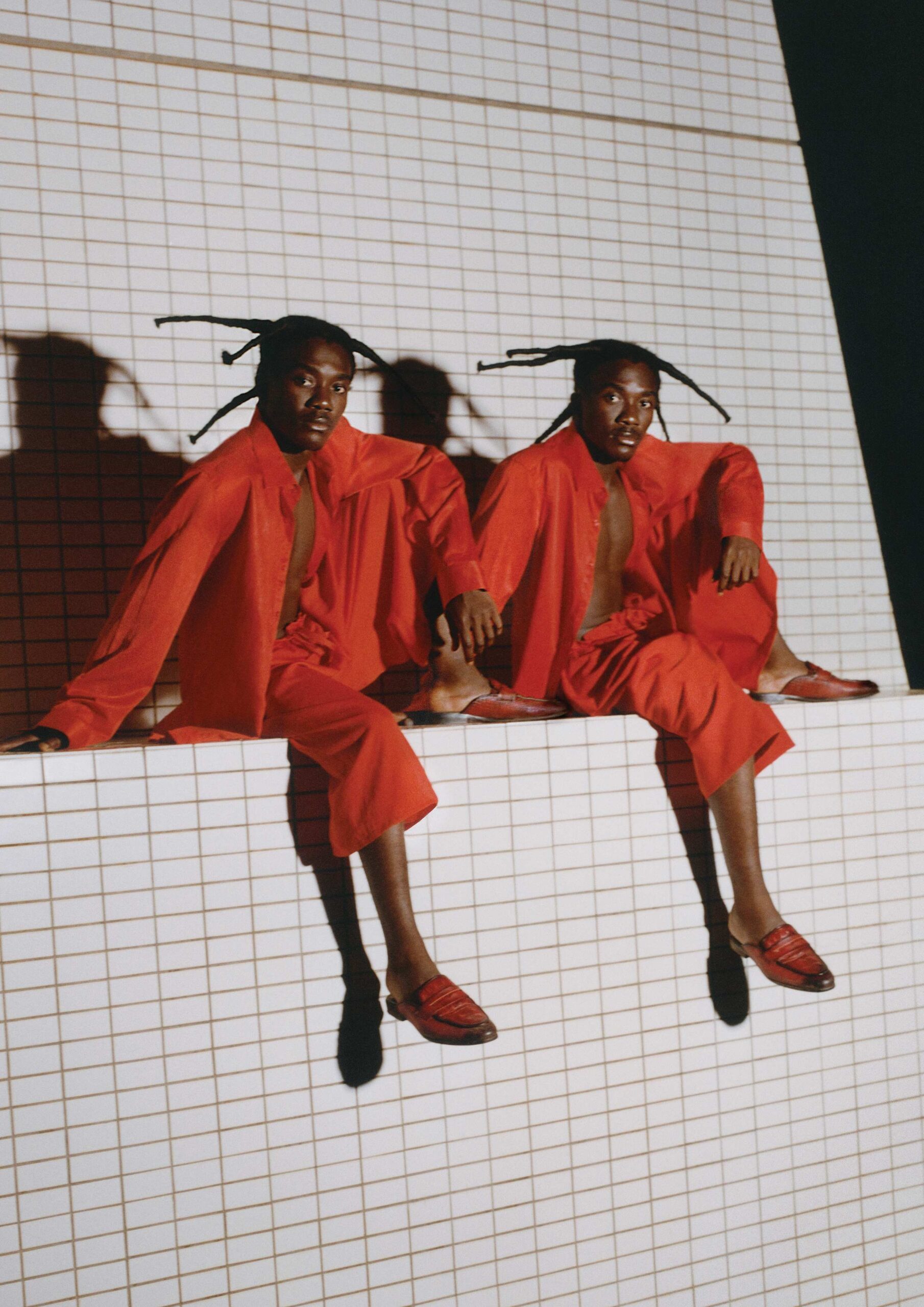 Two dancers in vibrant red outfits sitting on a white tiled structure against a deep red background, captured in a stylized, high-fashion pose for the Byredo Bal d’Afrique campaign.