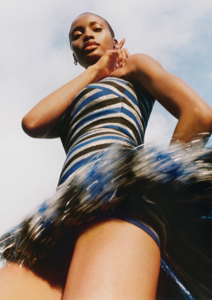 A low-angle fashion portrait of a model in a striped blue and gold Hertunba dress with a feathered skirt, posing against a clear blue sky for the Byredo Bal d’Afrique campaign.
