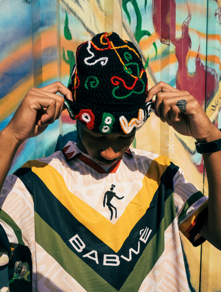 A man wearing a green, yellow, and white Zimbabwean-inspired football jersey and a matching patterned beanie, adjusting the hat while looking down