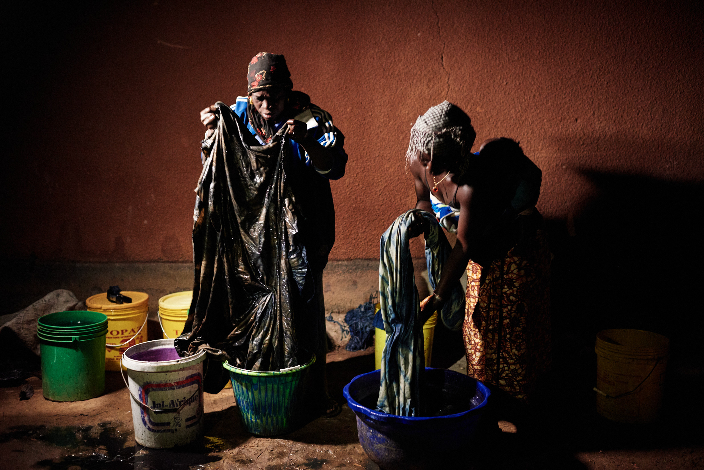Two women performing traditional indigo tie-dye, wringing dark dyed fabric over colorful basins in a workshop setting.
