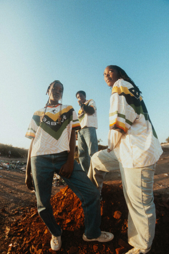 Out of Zimbabwe, a Streetwear Brand With Deeper Intentions 2 Three models standing outdoors wearing white Zimbabwean-inspired football jerseys with yellow and green accents, featuring the Babwê logo and chevron patterns.