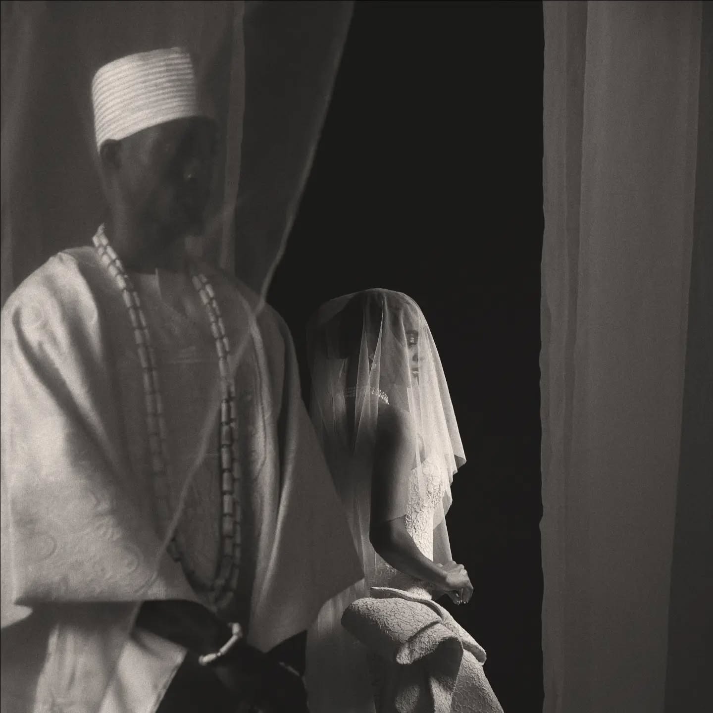 A black and white profile shot of a model wearing a structural 1930s-inspired hat and a layered traditional Nigerian outfit from the Ohlanna Bu Zikora collection.