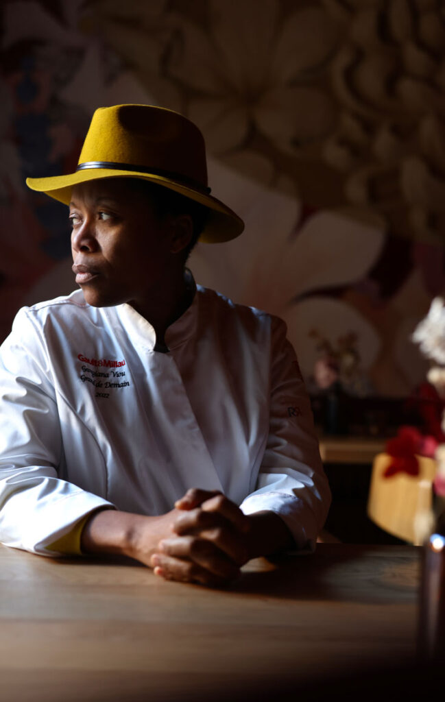 A portrait of Chef Georgiana Viou wearing a chef's uniform and a yellow hat, seated at L'Ami restaurant in Sofitel Cotonou Marina.