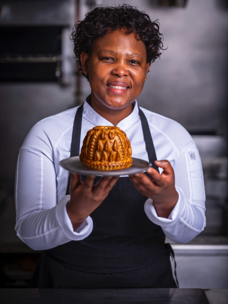 Chef Nokx Majozi wearing a white chef's uniform, smiling and holding one of her signature pies.