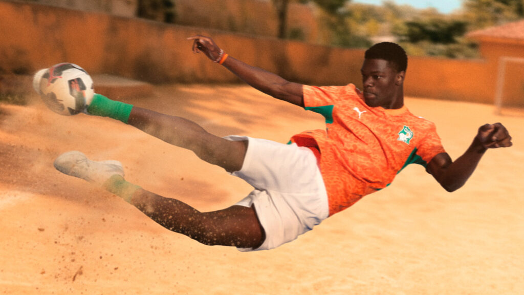Model wearing the orange and white PUMA Côte d'Ivoire national football team home kit for the 2026 World Cup sliding on a dirt pitch