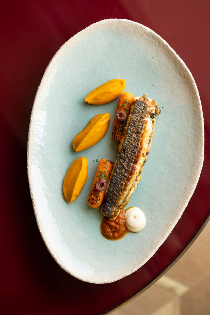 A top-down view of an elegantly plated dish on a pale blue plate, featuring small golden-brown fritters arranged with dollops of white sauce and delicate garnishes from L'Ami's kitchen.
