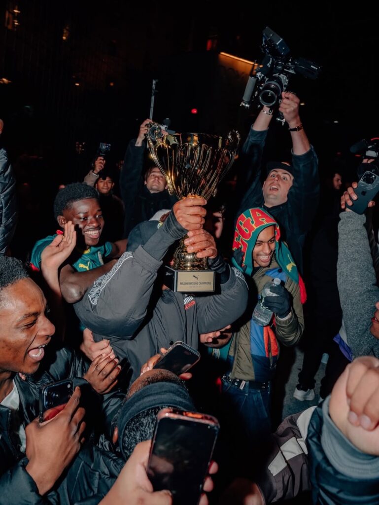 A man celebrating by holding up a golden boot trophy, surrounded by an excited crowd taking photos and videos with their smartphones.
