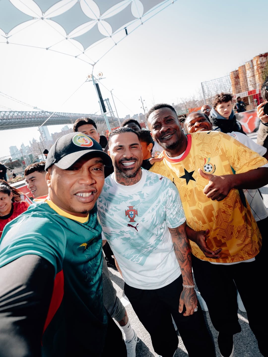 Asamoah Gyan wearing the yellow Ghana home jersey and El Hadji Diouf in the white Senegal jersey, smiling in front of a crowd at the PUMA 2026 World Cup kit launch event at Domino Park in Brooklyn.