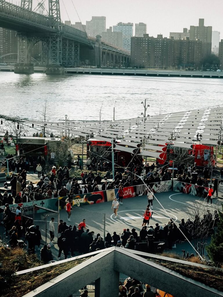 A wide shot of a large crowd gathered at the outdoor waterfront space in Domino Park, Brooklyn, for the PUMA 2026 World Cup kit launch event, with a suspension bridge in the background.