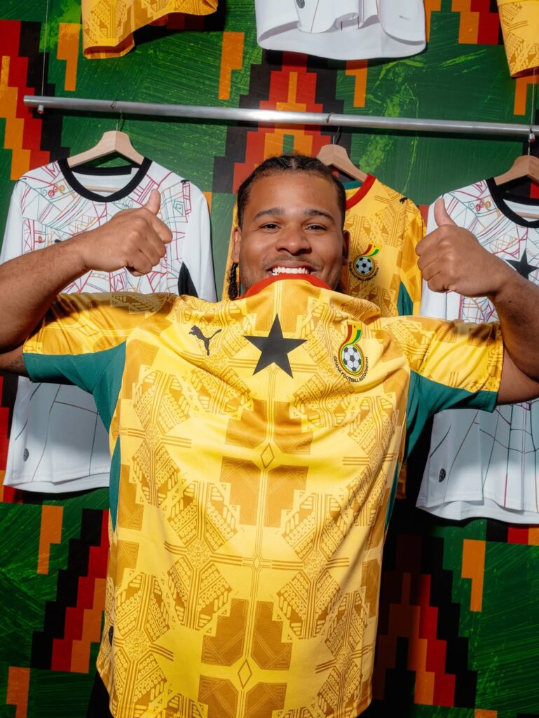 A man smiling and holding out the sleeves of a bright yellow football jersey featuring a black star in the center, standing indoors in front of a clothing rack.