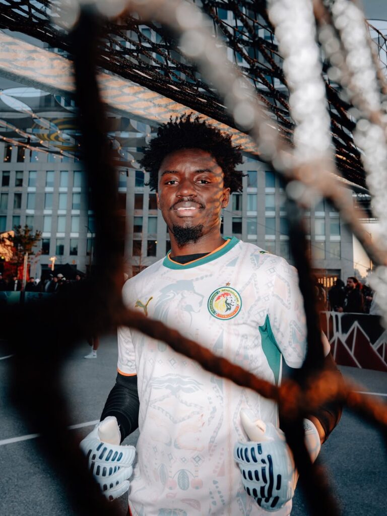 A smiling man wearing the white PUMA Senegal away jersey, standing outdoors behind a wire fence at the 2026 World Cup kit launch event.