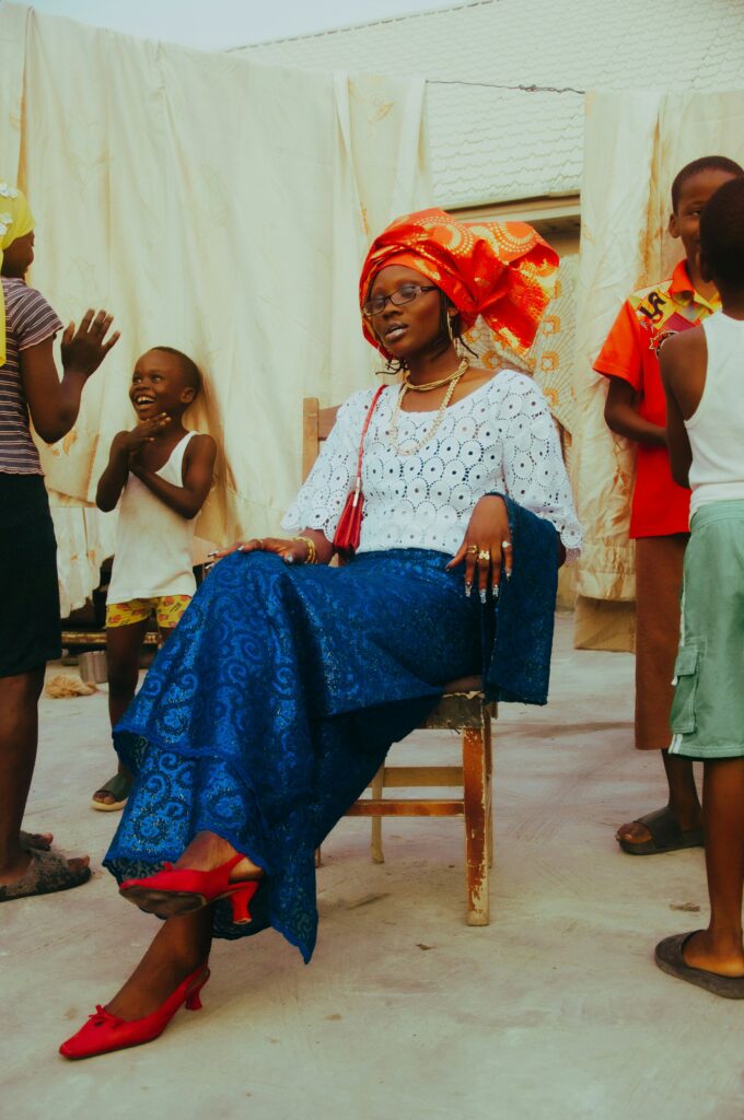 Why the Wrapper Remains West Africa's Undecolonized Garment 4 West African woman wearing a traditional blue lace iro and buba with a vibrant orange and red gele head tie, seated in a dignified portrait style.