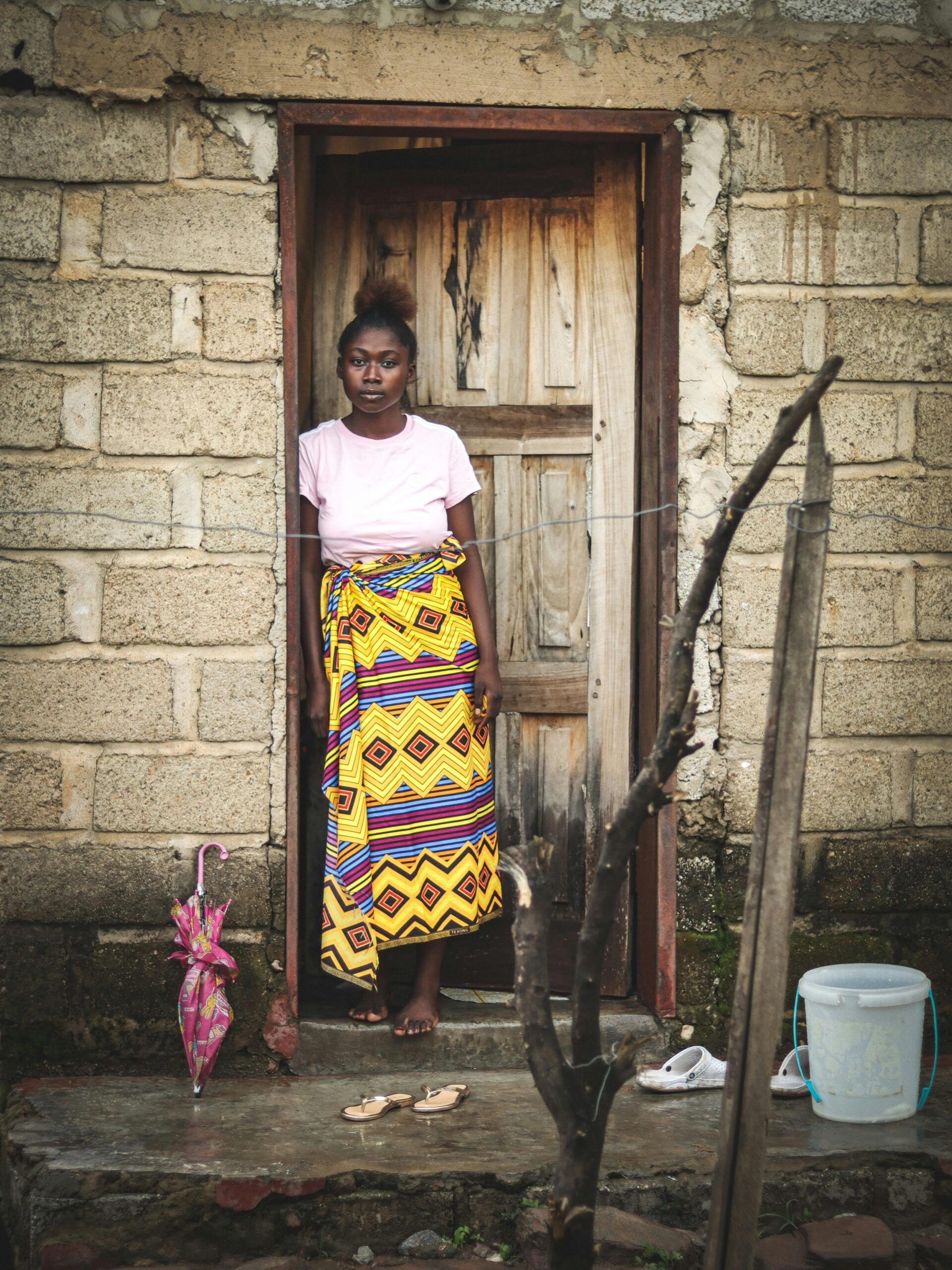 Young West African woman wearing a traditional vibrant yellow and purple patterned wrapper (lapa) standing in a rustic wooden doorway.