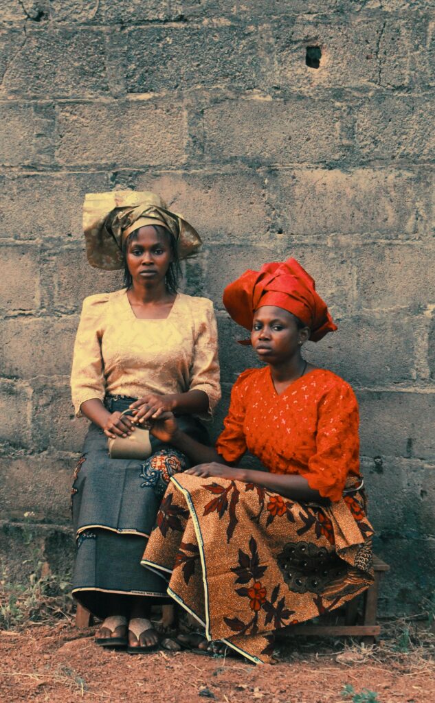 Why the Wrapper Remains West Africa's Undecolonized Garment 3 Two West African women wearing traditional George wrappers and matching lace blouses, featuring one woman in an ornate gold head tie and another in a vibrant red ensemble.