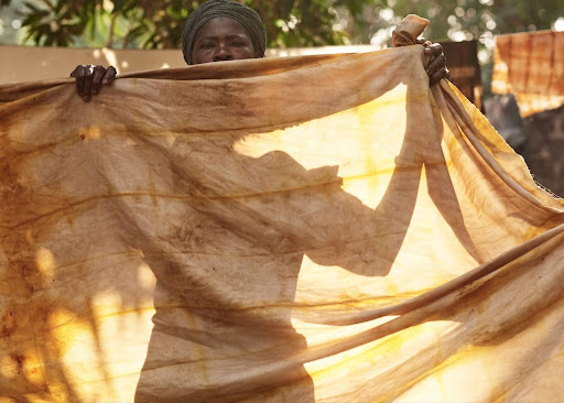 Material Literacy: The Knowledge in Cloth 5 A female artisan tie-dyer in Modiya, Guinea, holding up a large piece of naturally dyed cloth at a 2019 workshop led by Mariama Camara and Dr. Theanne Schiros.