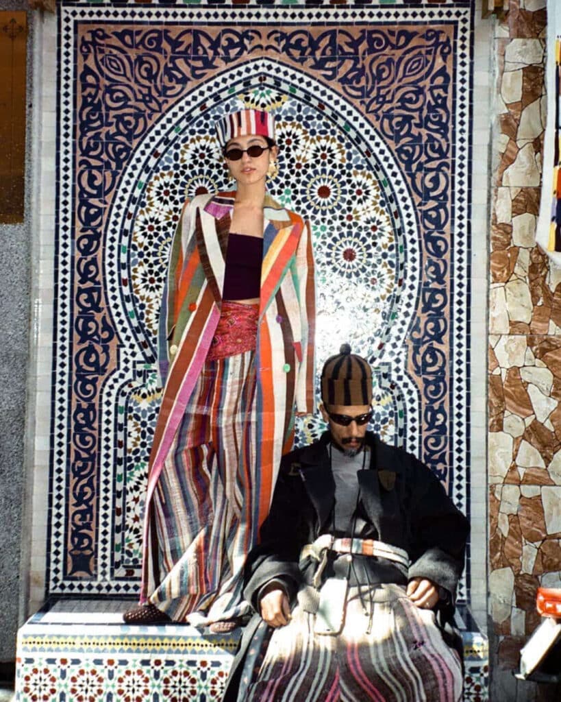 Woman in striped vintage blanket coat standing above Fahd Al Marsaoui seated below against a zellige mosaic fountain wall in Marrakech medina