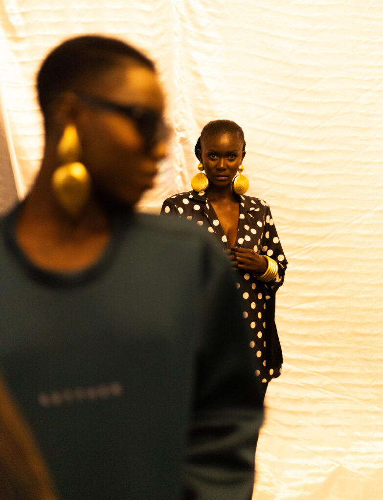 Market Entry Is Not a Mood Board 2 Backstage portrait of model in black and white polka dot blazer with oversized gold disc earrings and gold bangles, a second figure blurred in the foreground, shot in warm amber light against draped fabric.