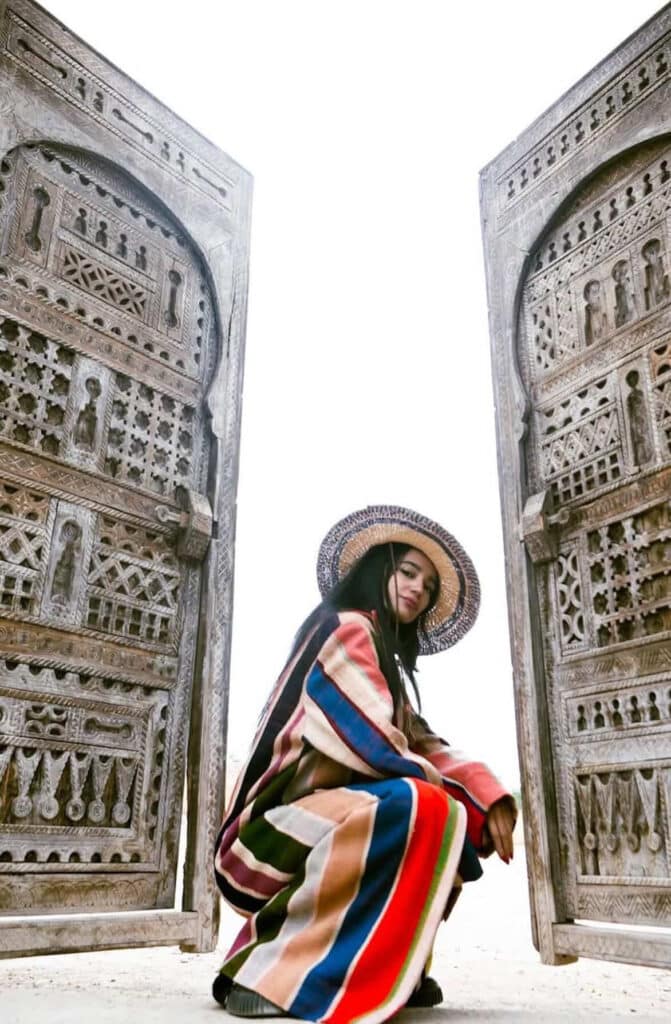 Woman in a multicolor striped blanket coat and woven straw hat crouching between two carved wooden Moroccan doors at Funky Cool Medina Marrakech
