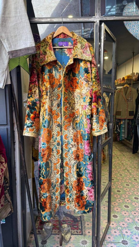 One-of-one floral velvet coat in orange blue and pink hanging in the doorway of Funky Cool Medina store in Marrakech with purple label visible
