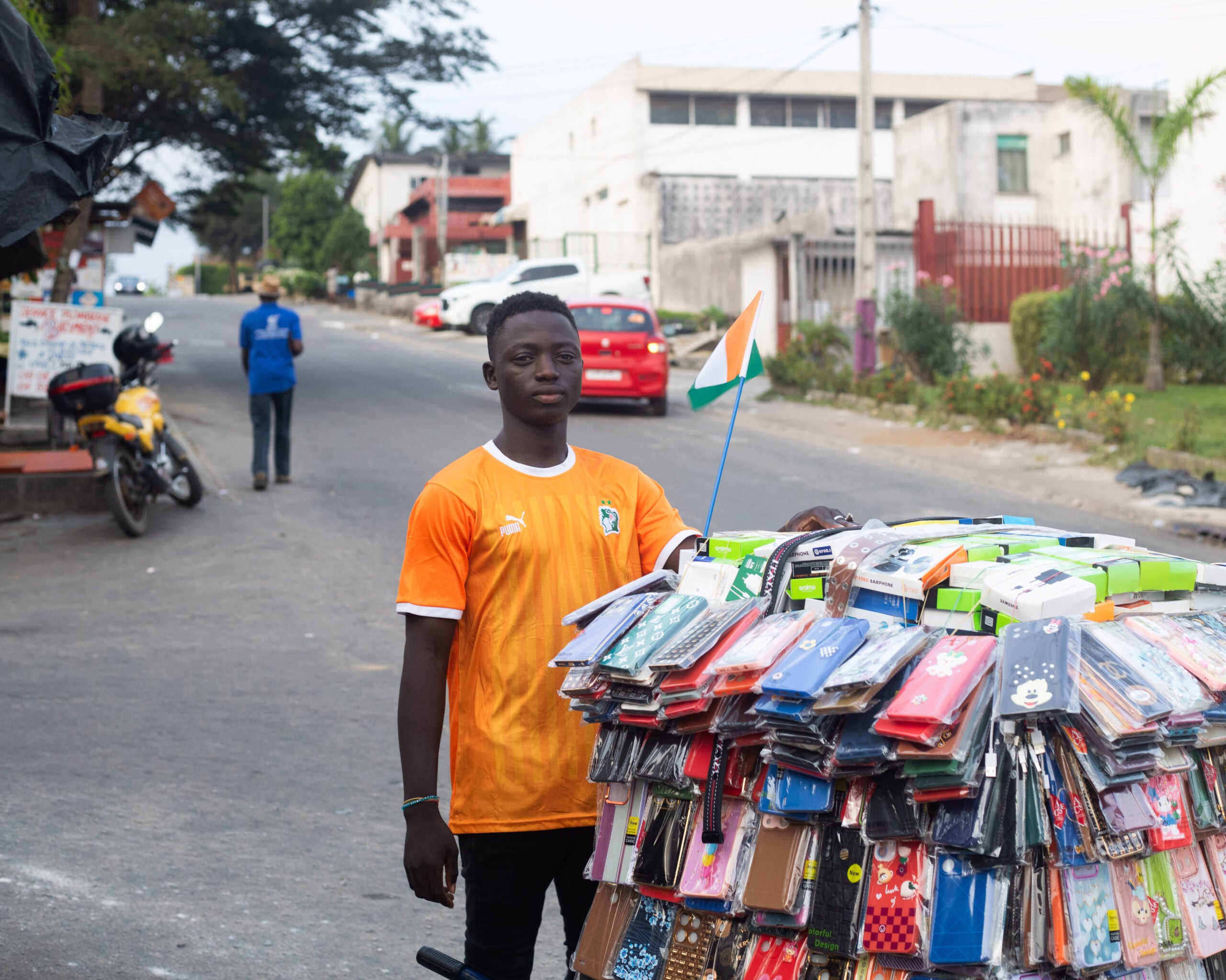 A street vendor in an orange shirt stands next to a roadside stall displaying colorful football jerseys in an urban setting.