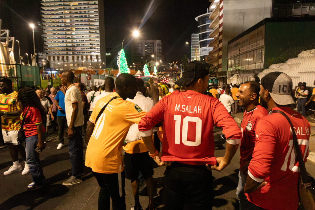 The Jersey Economy: Why Africa Is the Most Valuable Untapped Market in Sports Merchandise 5 African football fans wearing replica jerseys, including a red Liverpool Mo Salah shirt, gathered at a lively nighttime street viewing event.
