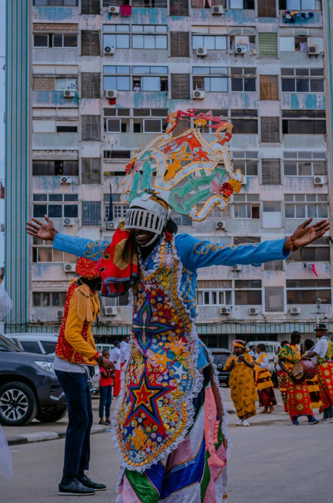 Before Rio, There Was Luanda 6 A person dressed in a bright blue and orange Carnival outfit with intricate headwear, posing with arms raised amid a street scene in Luanda, Angola.