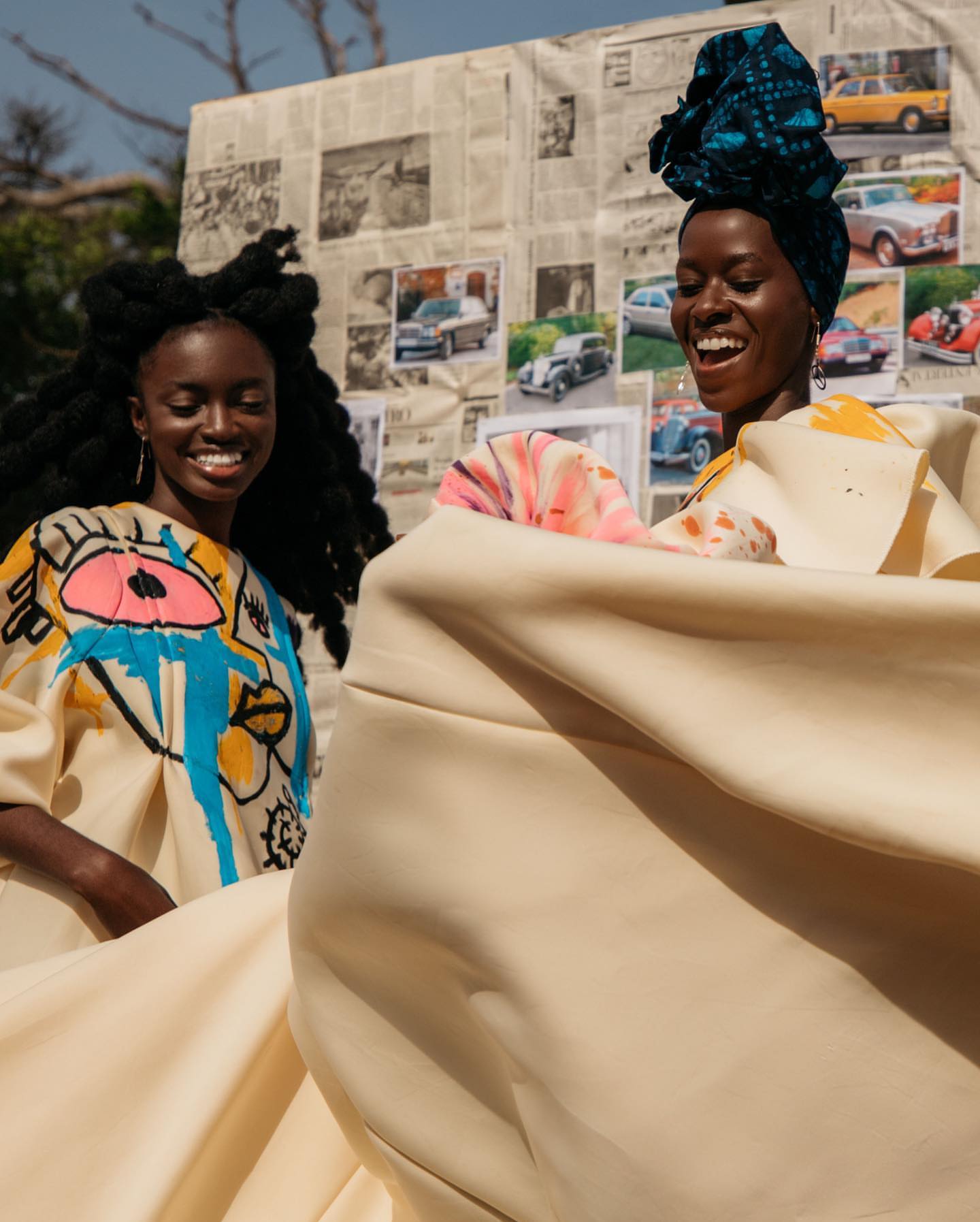 Two models in hand-painted garments laughing as cream fabric billows between them, photographed outdoors against a wall covered in newspaper clippings and vintage car photographs.