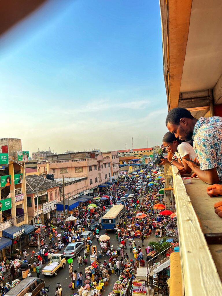 Market Entry Is Not a Mood Board 3 Aerial view of a West African market from a balcony, showing hundreds of traders under colorful umbrellas filling the street between commercial buildings, with onlookers photographing the scene from above.