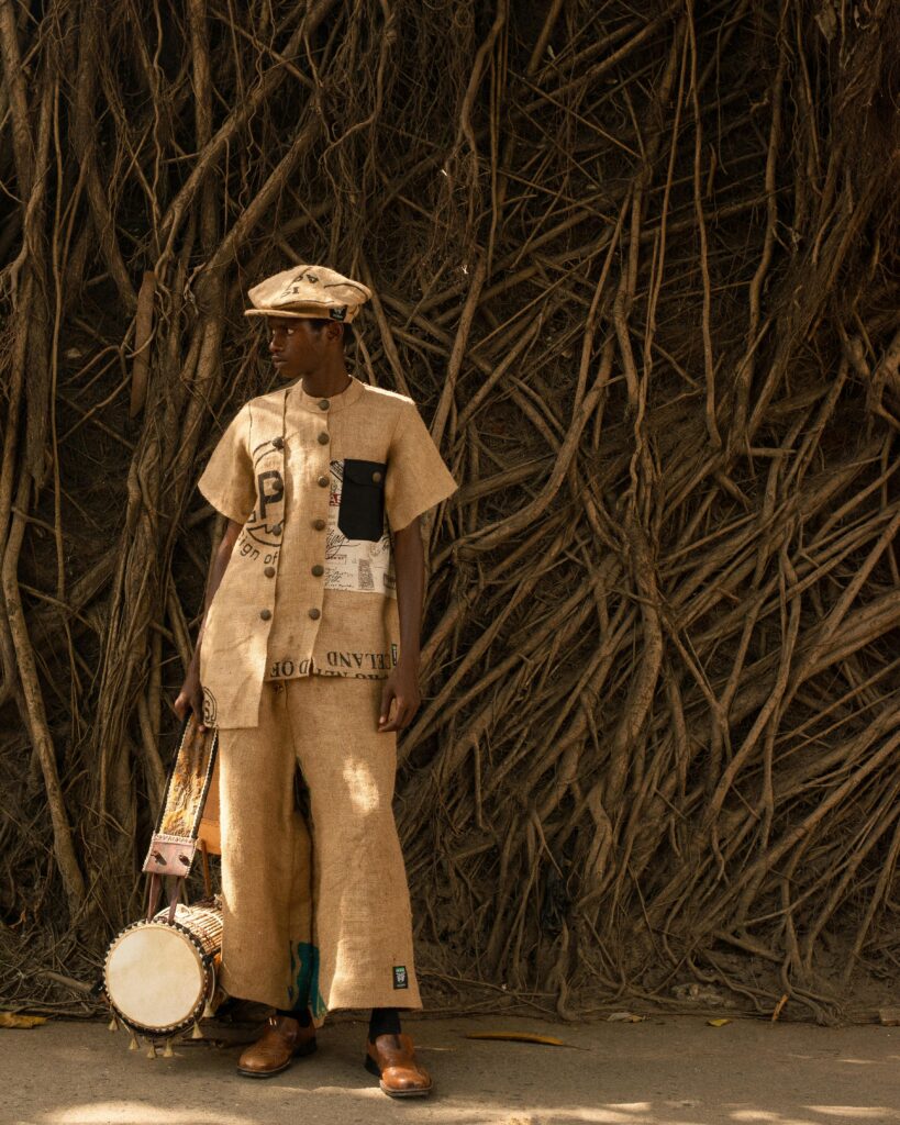 Market Entry Is Not a Mood Board 4 Model in full burlap suit with visible trade markings and brass buttons, wearing a matching cap and holding a traditional drum, standing against a wall of dense aerial tree roots in natural light.
