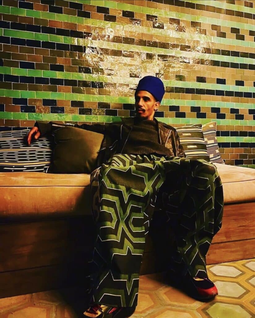 Funky Fahd Will See You Now 2 Funky Cool Medina founder Fahd Al Marsaoui seated on a sofa in green geometric pants and blue cap against green zellige tiles in Marrakech