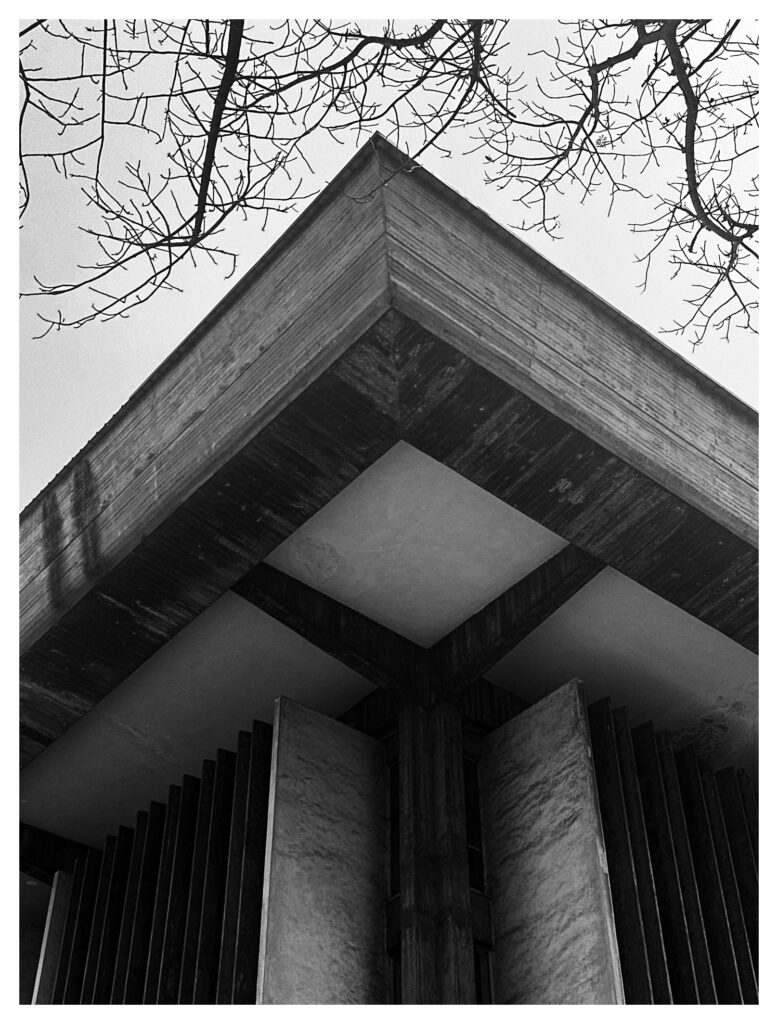 Upward view of a raw concrete corner overhang at the University of Lagos framed by bare tree branches, photographed in black and white by Uche Ibemere.