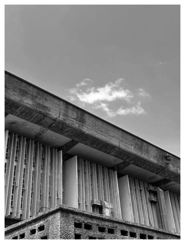 Wide view of a brutalist building at the University of Lagos showing vertical concrete fins beneath a dramatic horizontal overhang, with a textured perimeter wall in the foreground.