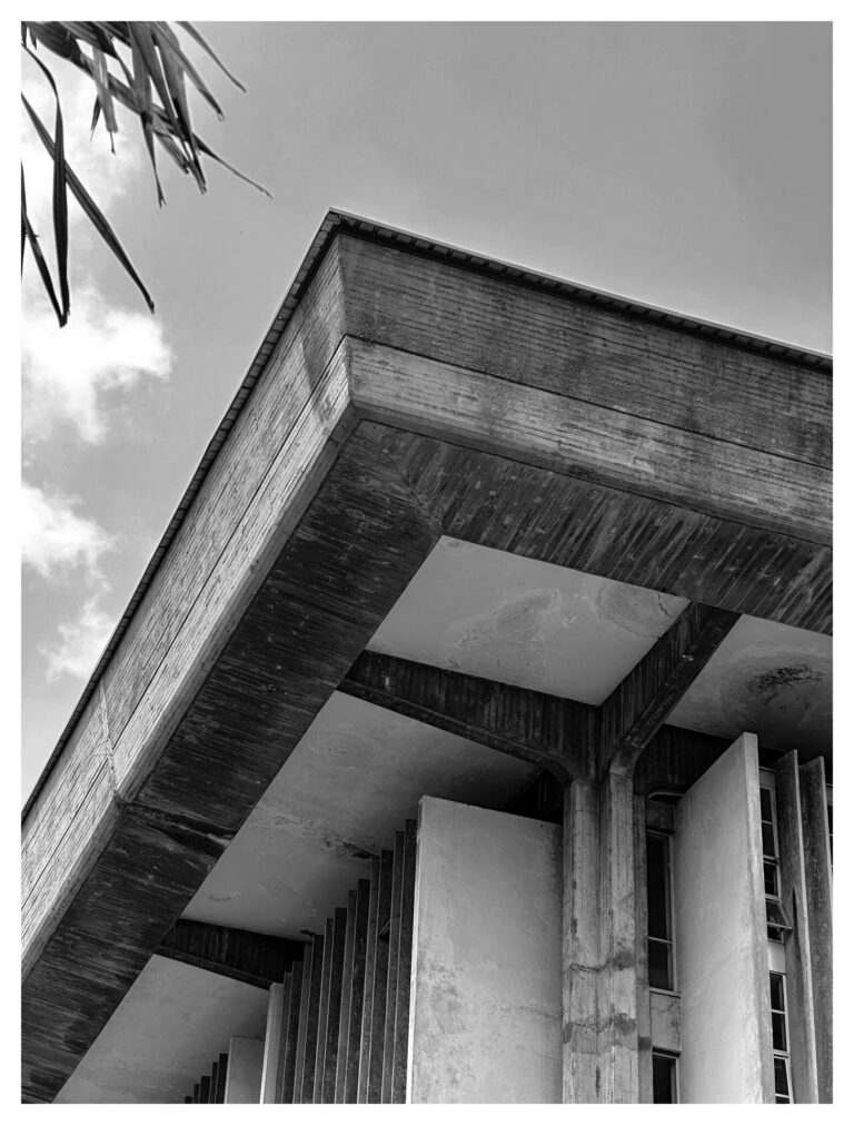 Upward view of a massive concrete cantilevered overhang supported by columns at the University of Lagos, with vertical fins and a palm frond visible against the sky.