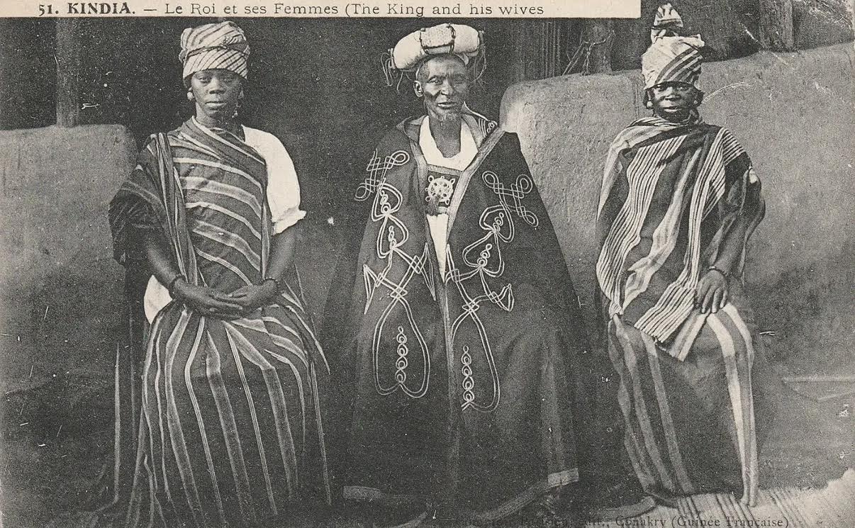 Black and white 1910 portrait of the King of Kindia and his wives dressed in traditional Basse-Guinée textiles, highlighting historical tie-dye and handwoven embroidery.