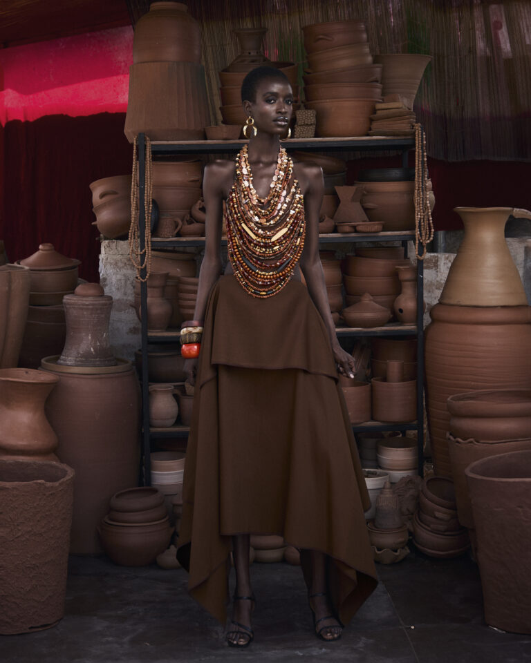 Model wearing a brown tiered skirt and intricate multi-layered clay bead statement necklace from Hertunba's Akaoru collection, standing among traditional terracotta pots.