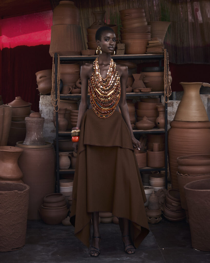Model wearing a brown tiered skirt and intricate multi-layered clay bead statement necklace from Hertunba's Akaoru collection, standing among traditional terracotta pots.