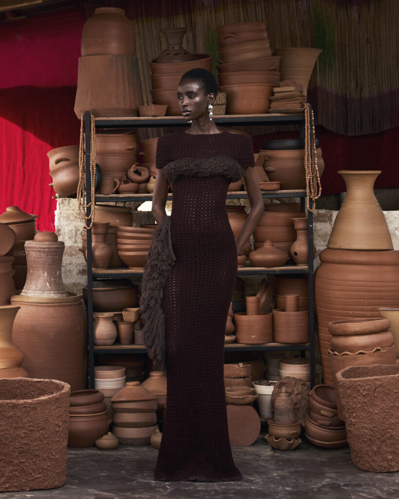 Hertunba's 'Akaoru' Collection Is a Lesson in What Craftsmanship Actually Costs 9 Model wearing a floor-length, textured dark brown gown from Hertunba's Akaoru collection, standing in a studio surrounded by shelves of traditional terracotta vessels.
