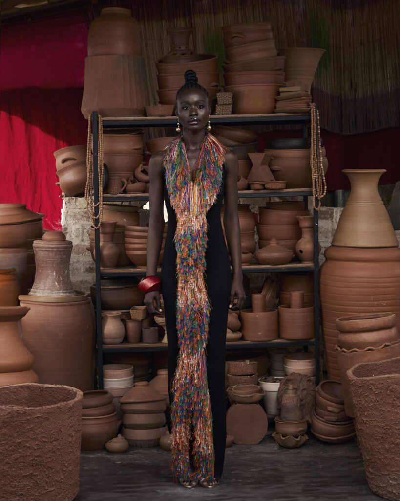 Hertunba's 'Akaoru' Collection Is a Lesson in What Craftsmanship Actually Costs 8 Model wearing a vividly colored, plunging halter-neck dress with intricate vertical patterns from Hertunba's Akaoru collection, standing among traditional terracotta pots in a studio.