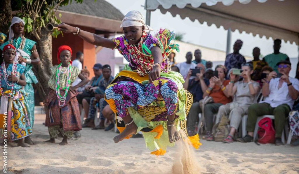 Cotonou Knows Exactly What It's Doing 5 Young Vodun practitioner mid-jump during a ceremony in Ouidah, Benin, wearing cowrie shells and traditional dress as an audience watches.