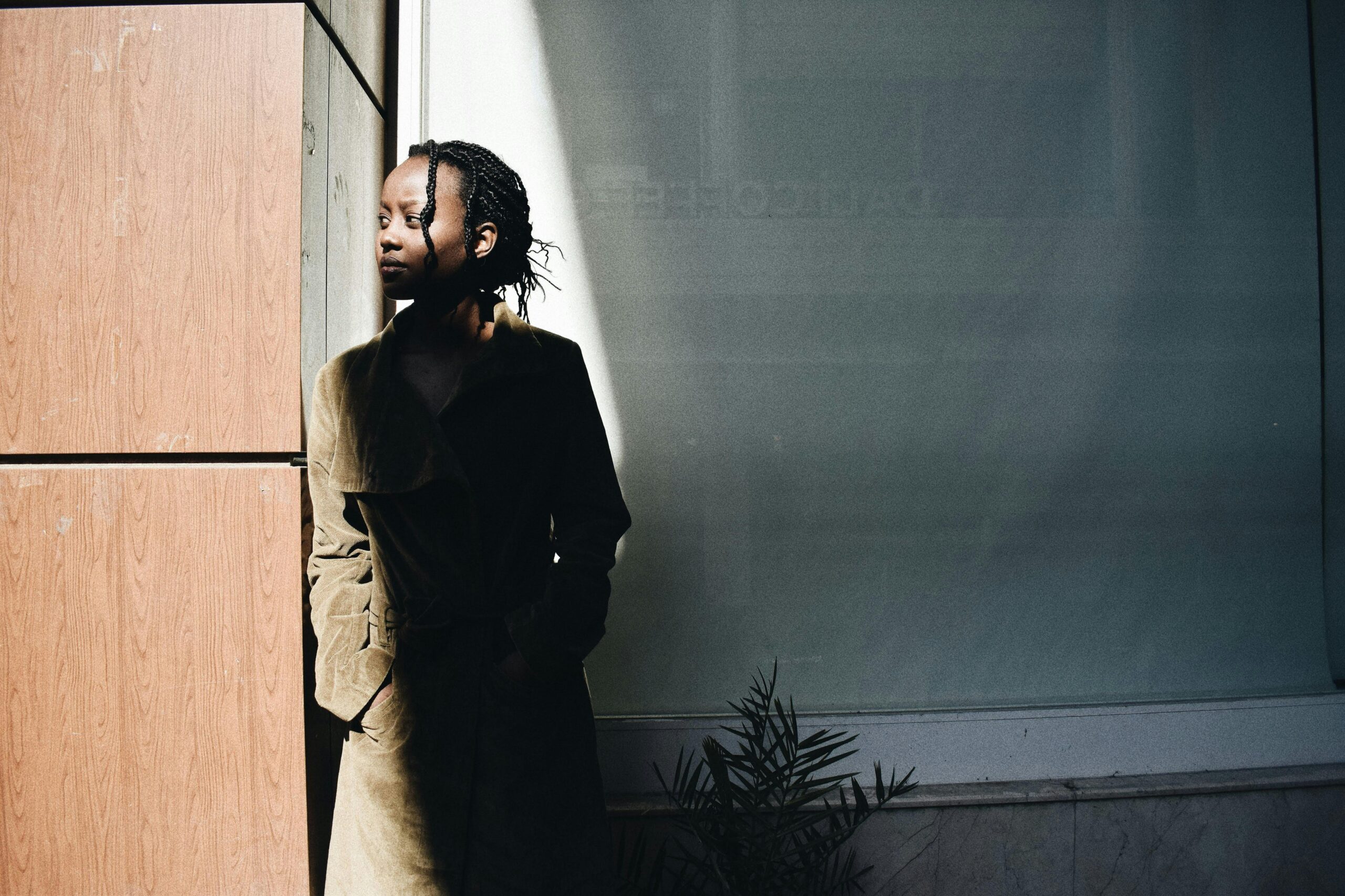 A woman in a dark coat standing against a wall with strong sunlight and shadow patterns.