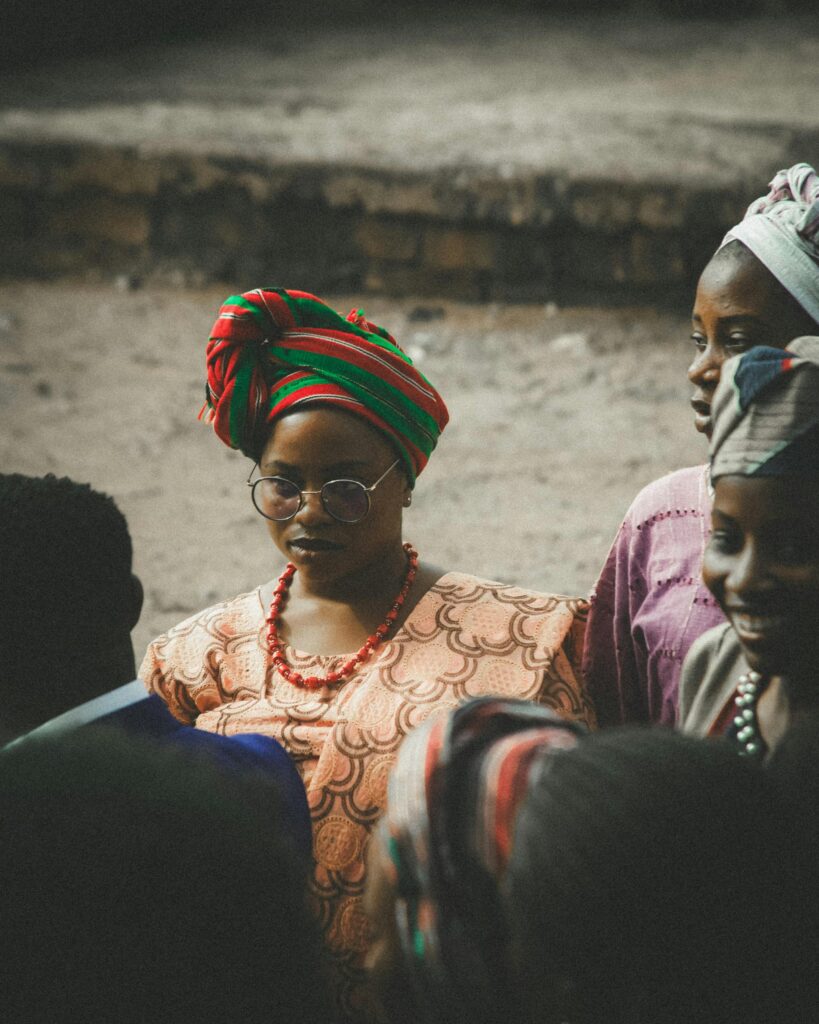 Woman wearing a colorful headwrap and round glasses seated among others in a communal gathering