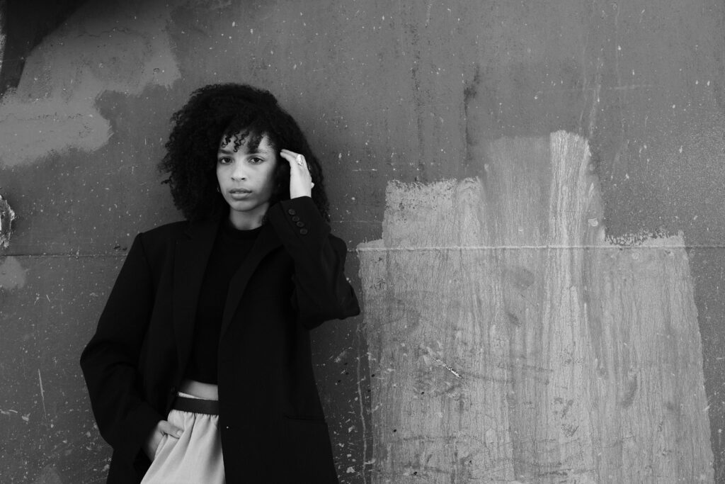 A black and white portrait of a woman in a dark coat standing against a textured wall.