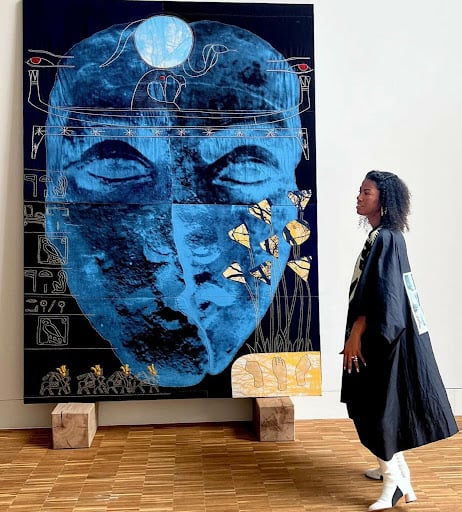 Ghanaian-German artist Zohra Opoku standing in her studio next to a monumental blue stitched textile mask, highlighting contemporary African art and memory.