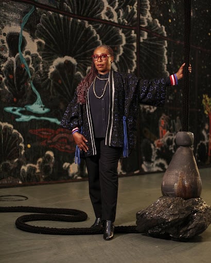Woven Stories: 9 African Artists Shaping Contemporary Narratives Through Textiles 8 Contemporary African artist Otobong Nkanga standing in front of a monumental woven tapestry and mixed-media installation, highlighting material storytelling and global economies.