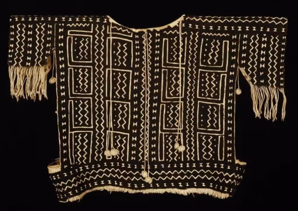 Understanding Bògòlanfini: The Malian Textile Written in Mud and Cotton 24 A 20th-century Bògòlanfini tunic from Tenenkou, Mali, with cream geometric motifs on a dark mud-dyed ground, fringed sleeves, and bell tassels, photographed against a black background. Collection of the Minneapolis Institute of Art.