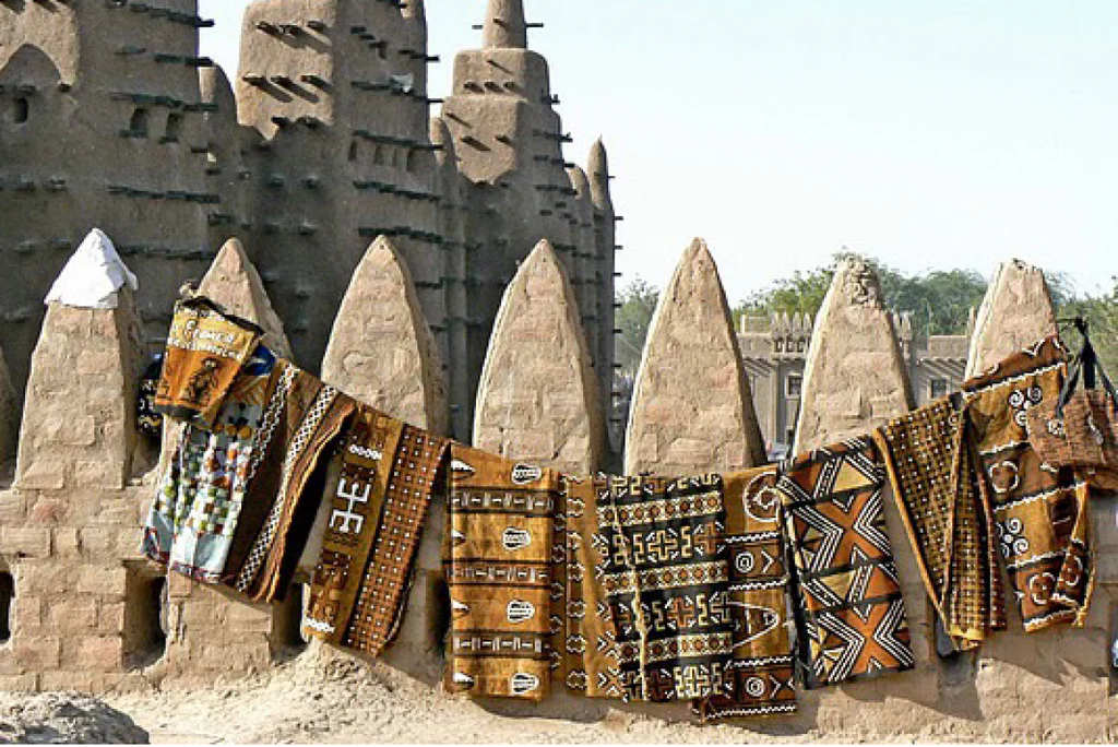 Multiple bògòlanfini cloths in earth tones hang horizontally against the conical mud-brick pillars of the Great Mosque of Djenné, Mali, under bright sunlight.