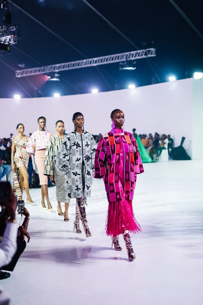 After the Applause: Heritage Textiles and the Economics of the Runway Moment 59 Five Black women models walk a runway in a curved white-walled venue, wearing handwoven heritage textiles tailored as contemporary luxury garments. The model in the foreground wears a pink and red kente coat with a long fringed skirt and metallic boots. Beside her, a model in a grey and black patchwork coat with appliqué motifs. Behind them, three more models in striped, geometric, and embellished handwoven pieces. Lagos runway lighting, audience visible at the edges.
