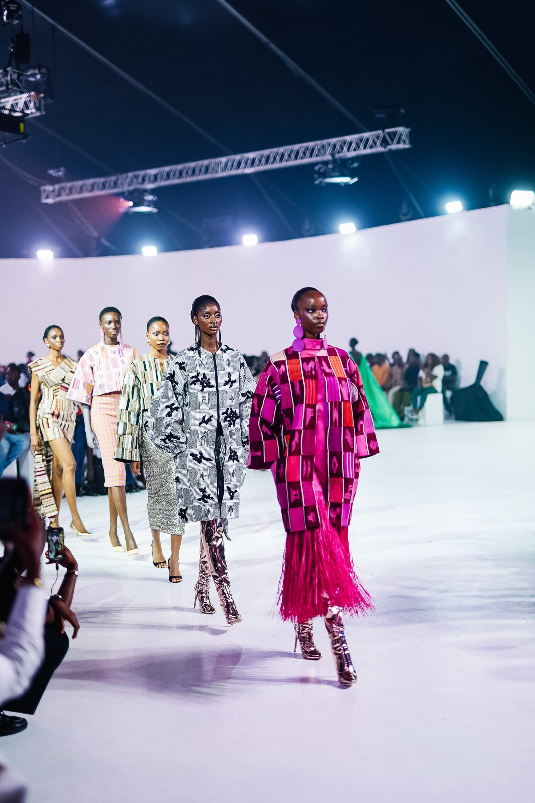 Five Black women models walk a runway in a curved white-walled venue, wearing handwoven heritage textiles tailored as contemporary luxury garments. The model in the foreground wears a pink and red kente coat with a long fringed skirt and metallic boots. Beside her, a model in a grey and black patchwork coat with appliqué motifs. Behind them, three more models in striped, geometric, and embellished handwoven pieces. Lagos runway lighting, audience visible at the edges.