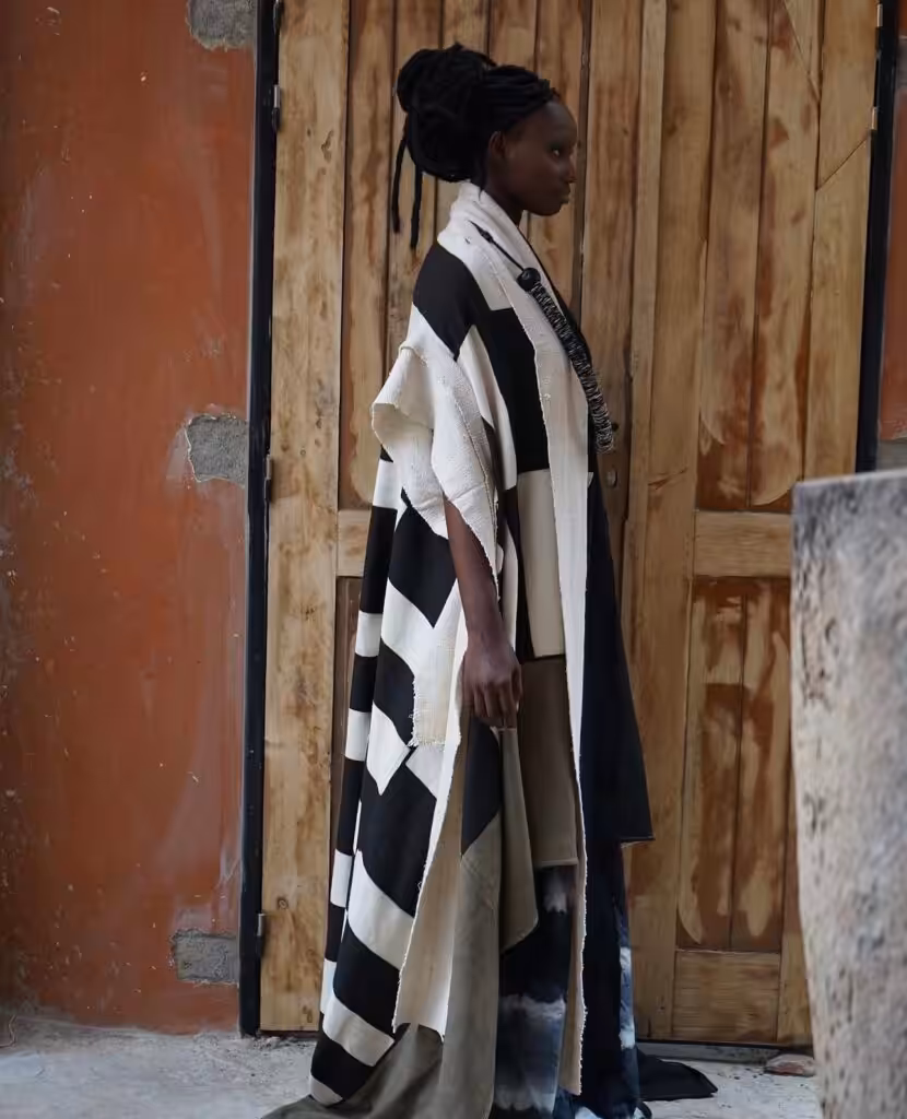 A model in a flowing patchwork garment composed of bògòlanfini, indigo, and natural cream and black panels, photographed in profile against a weathered orange wall and wooden door, from Awa Meïté's Bogolan collection paying homage to Chris Seydou.